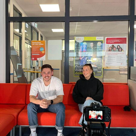 Zwei Personen sitzen auf einem roten Sofa und werden von einer Kamera gefilmt, als ob sie ein Interview führen.
