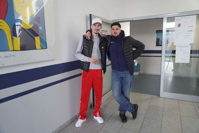 Zwei Männer posieren lässig in einem Flur vor einem bunten Bild, einer trägt rote Hosen und eine weiße Mütze.