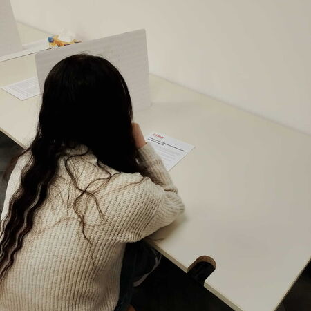 Ein Mädchen mit langen Haaren sitzt an einem Tisch, konzentriert auf ein Blatt Papier vor sich.