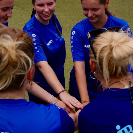 Eine Gruppe junger Frauen in blauen Trikots legt die Hände beim Teamspiel auf einem Sportplatz zusammen.