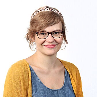 Eine Frau mit Brille lächelt in die Kamera und trägt ein gelbes Oberteil sowie ein Haarband, vor einem weißen Hintergrund.
