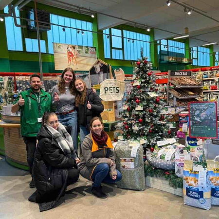 Eine Gruppe von Menschen posiert fröhlich vor einem geschmückten Weihnachtsbaum in einem gut sortierten Tierbedarfsgeschäft.