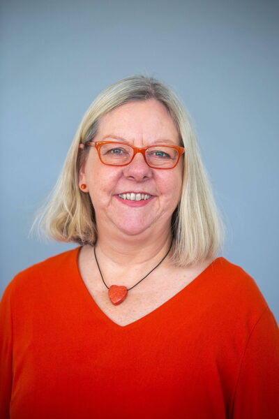 Eine lächelnde Frau mit blonden Haaren trägt eine orange Brille und ein passendes Oberteil vor einem grauen Hintergrund.