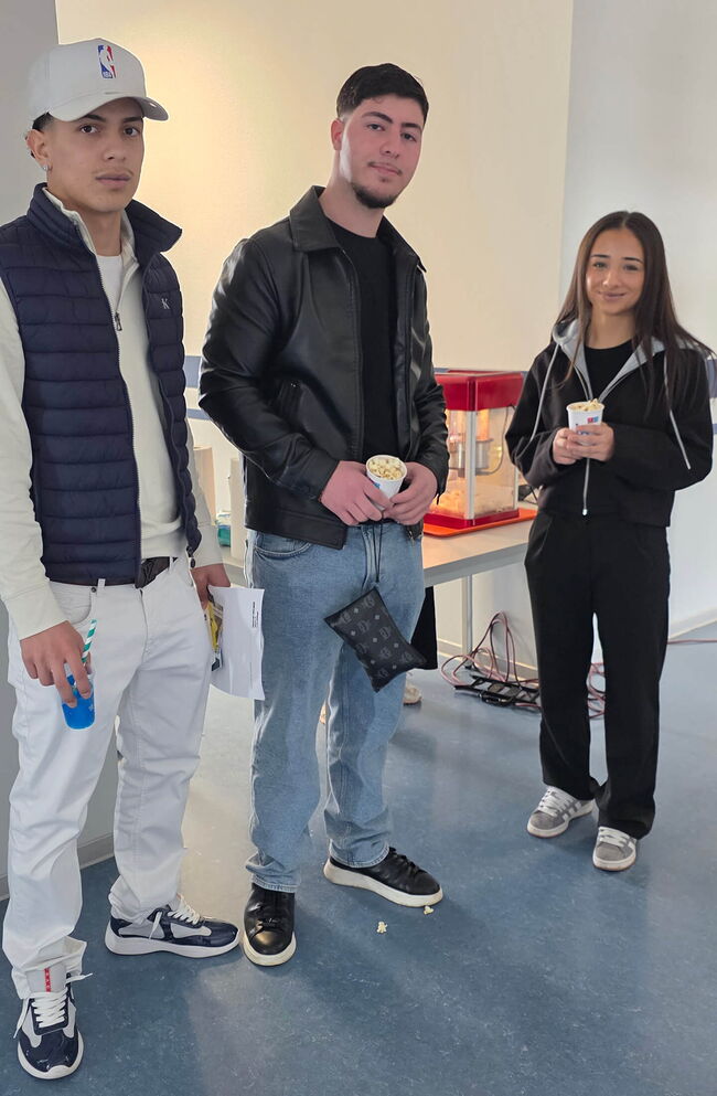 Drei Schüler stehen in einem Raum und halten Snacks, während im Hintergrund eine Popcornmaschine zu sehen ist.