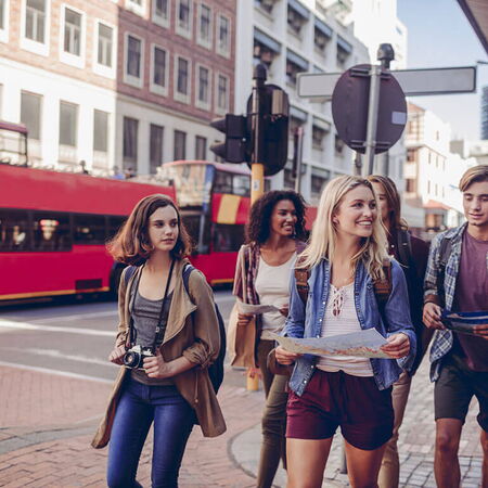 Eine Gruppe junger Menschen erkundet mit Karten eine belebte Stadt, während ein roter Doppeldeckerbus vorbeifährt.