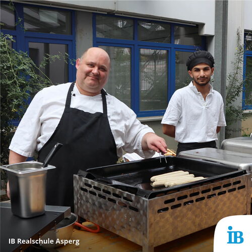 Ein Koch und ein Assistent bereiten beim Schulfest Würstchen auf einem Grill im Freien zu.