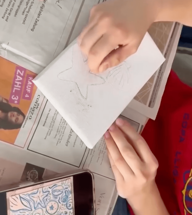Ein Kind zeichnet mit einem Bleistift auf einem Blatt Papier einen Stern, während es auf einer Zeitung sitzt.
