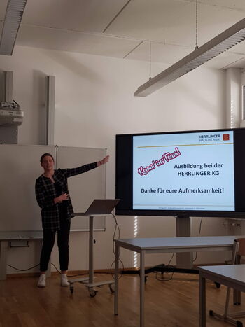 Eine Person präsentiert in einem Raum vor einem Bildschirm, der Informationen zur Ausbildung bei HERRLINGER KG zeigt.