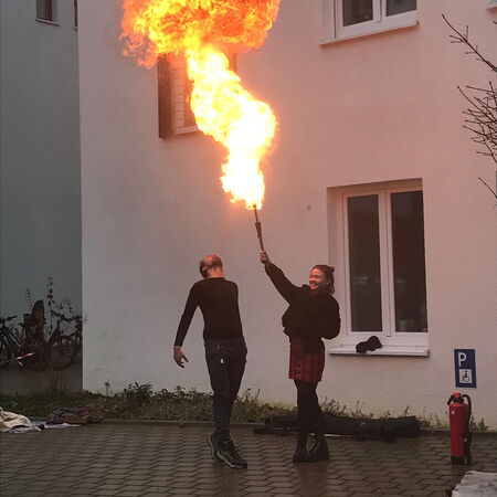 Eine Frau hält eine Fackel, aus der eine große Flamme schießt, während ein Mann daneben überrascht zurückweicht.