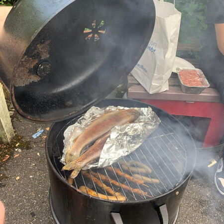 Frische Forellen werden in Alufolie auf einem runden Grill im Freien geräuchert, während Rauch aufsteigt.