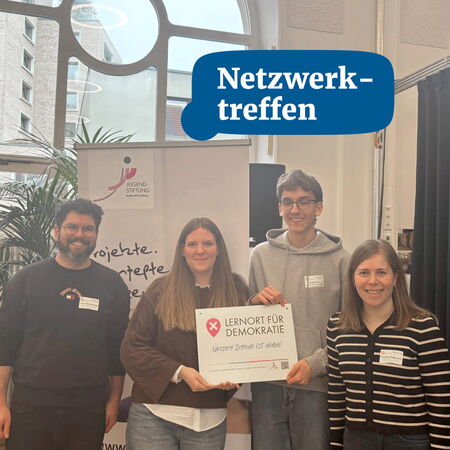 Vier Personen stehen lächelnd vor einer Wand mit dem Banner "Lernort für Demokratie" bei einem Netzwerk-Treffen.