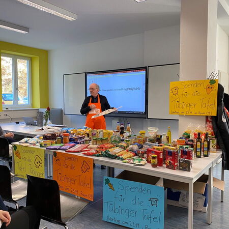 Ein Mann in einer orangefarbenen Schürze präsentiert gesammelte Lebensmittelspenden für die Tübinger Tafel in einem Klassenzimmer.