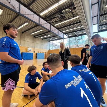 Eine Gruppe junger Männer in blauen Trikots sitzt in einer Turnhalle und hört einer stehenden Frau aufmerksam zu.