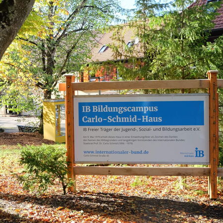 Ein Schild des IB Bildungscampus Carlo-Schmid-Haus steht zwischen herbstlichen Bäumen auf einem sonnigen Gelände.