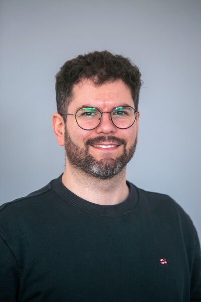 Ein lächelnder Mann mit Brille und Bart trägt ein schwarzes Oberteil vor einem neutralen Hintergrund.
