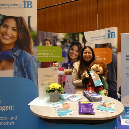 Eine Frau steht lächelnd an einem Stand des Bildungscampus Gesundheit & Soziales mit Infobroschüren und einem Stofftier.