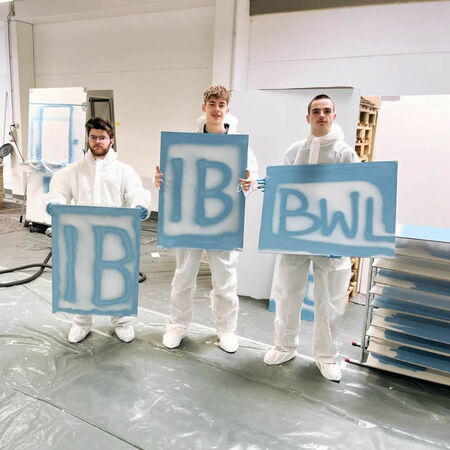 Drei Personen in Schutzanzügen halten Schilder mit den Buchstaben "IB" und "BWL" in einer Werkstatt, die Jobrotation darstellt.