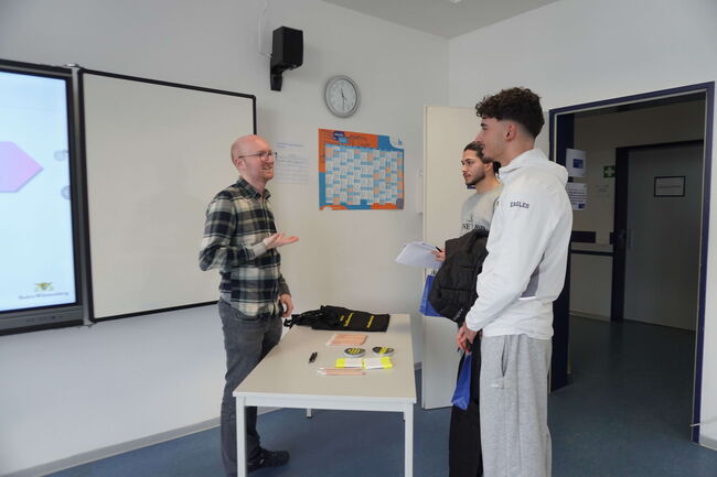 Ein Lehrer unterhält sich mit zwei Schülern in einem Klassenzimmer neben einer Tafel und einem Tisch.