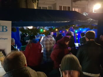 Eine Gruppe von Menschen versammelt sich unter einem festlich geschmückten Stand bei Abenddämmerung auf einem Weihnachtsmarkt.