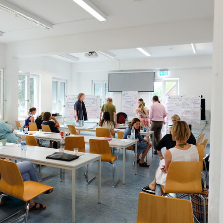 Teilnehmer in einem Seminarraum diskutieren intensiv vor Flipcharts, während andere an Tischen sitzen und Notizen machen.