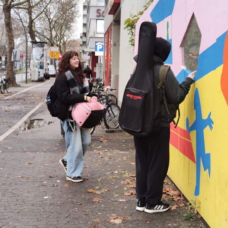 Zwei Personen mit Musikinstrumenten-Taschen stehen vor einer bunt bemalten Wand in einer städtischen Umgebung.