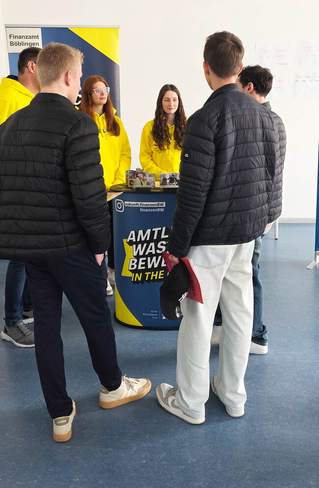 Eine Gruppe junger Menschen steht am Stand des Finanzamts Böblingen und spricht mit Mitarbeitern in gelben Jacken.