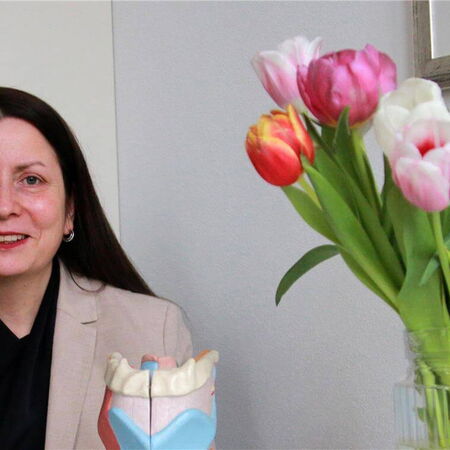 Eine Frau sitzt lächelnd neben einem Kehlkopfmodell, mit einem Strauß bunter Tulpen im Vordergrund.