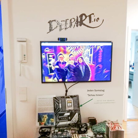 Ein Fernseher zeigt ein bunt verfremdetes Bild von zwei Personen, während darunter Computerteile auf einem Tisch liegen.