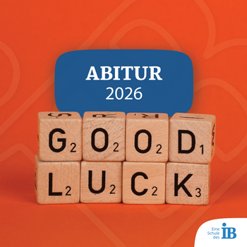 Holzwürfel zeigen die Botschaft "GOOD LUCK" und das Abitur-Jahr 2026 auf einem leuchtend orangefarbenen Hintergrund.