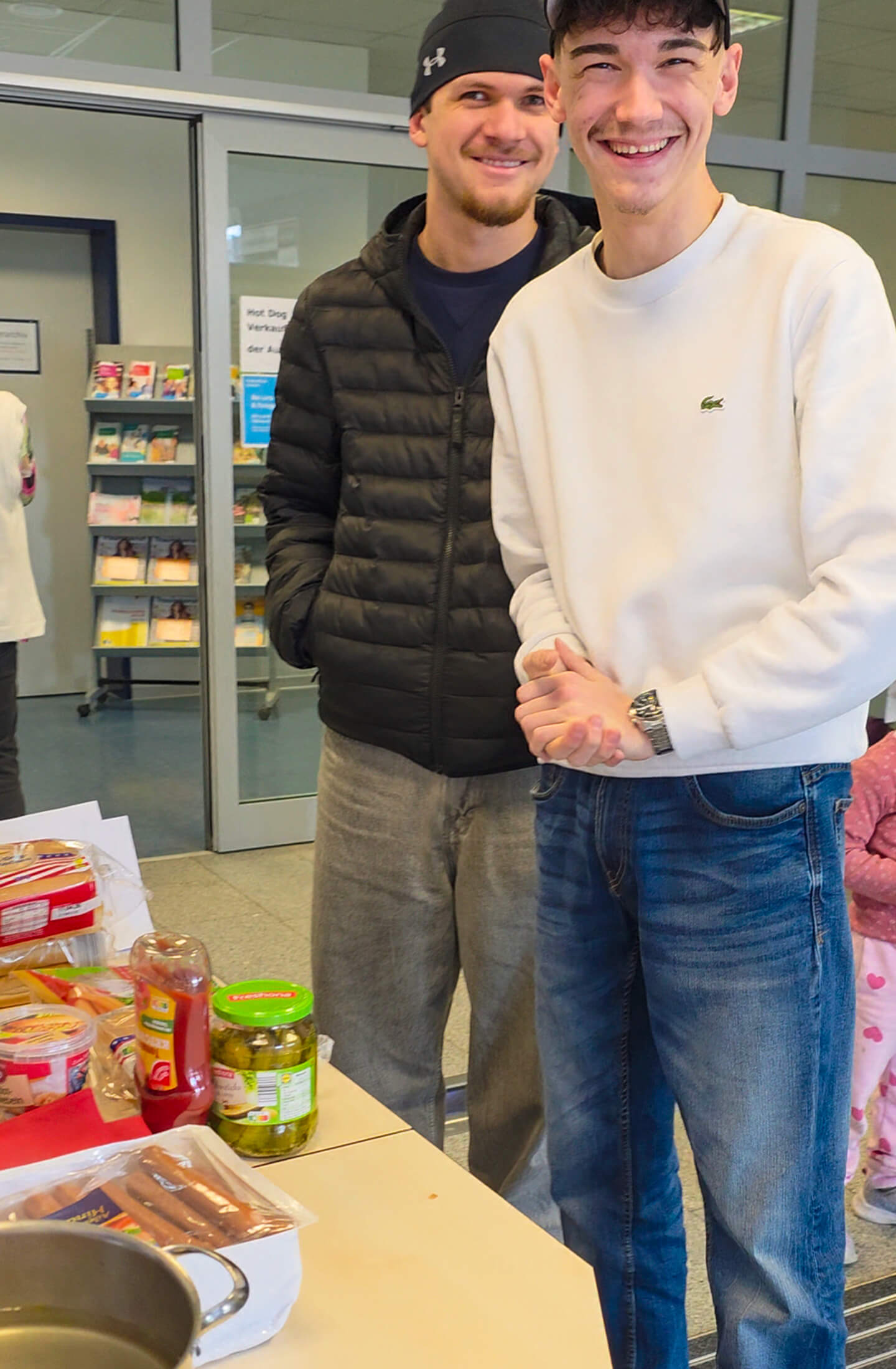 Zwei junge Männer stehen lächelnd an einem Tisch mit Hot-Dog-Zutaten und genießen den Tag der offenen Tür.