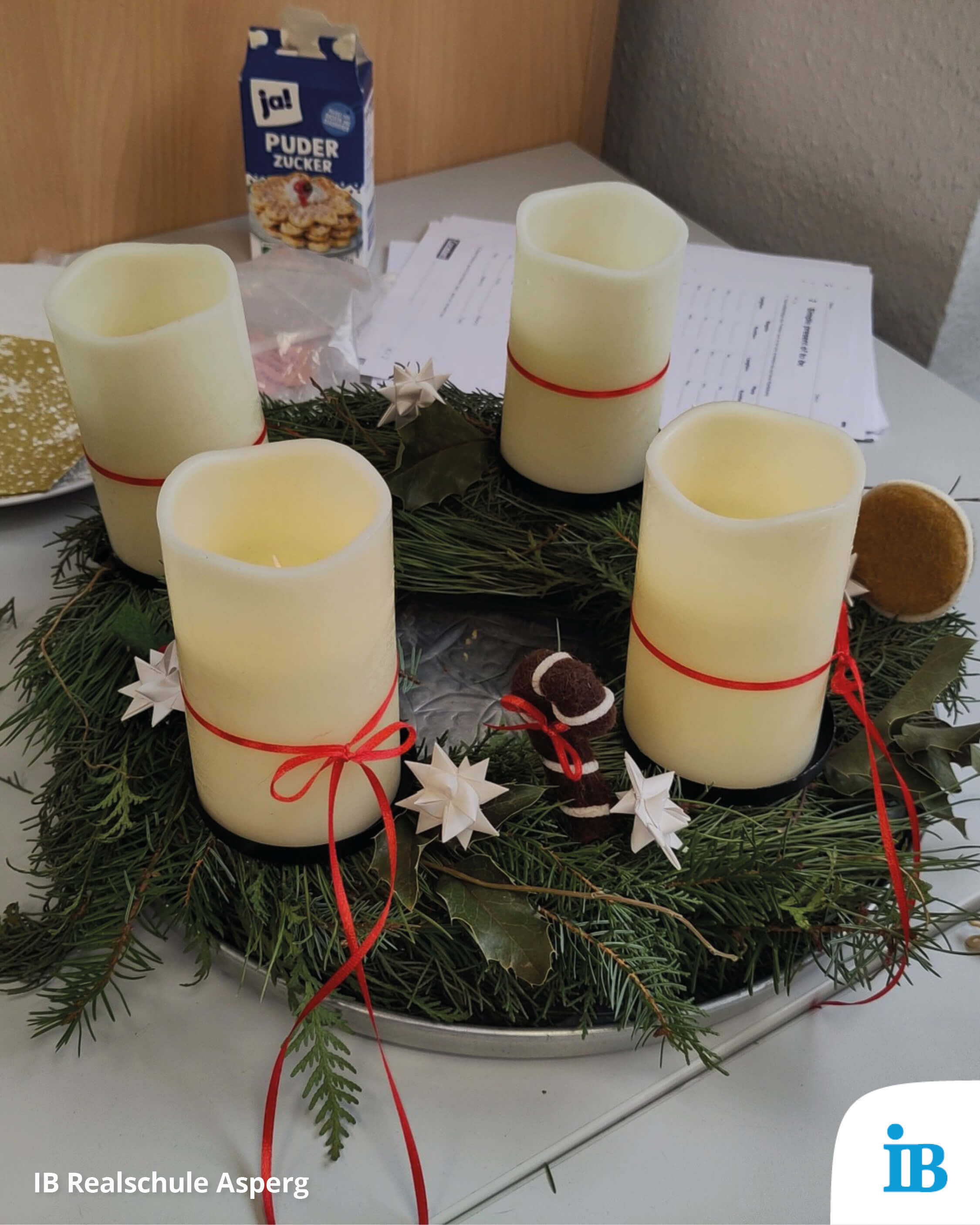 Ein Adventskranz mit vier Kerzen und roten Schleifen steht neben einer Packung Puderzucker auf einem Tisch.
