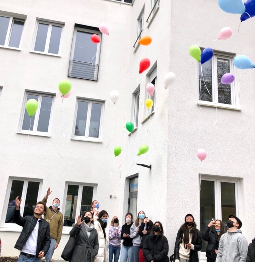 Menschen lassen bunte Luftballons vor einem weißen Gebäude steigen und schauen freudig in den Himmel.
