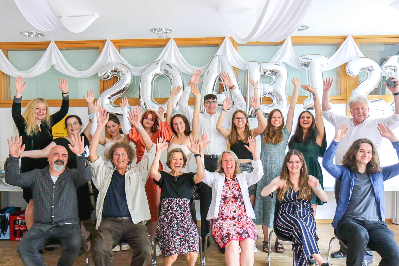 Eine fröhliche Gruppe von Menschen feiert den Abiturjahrgang 2021 mit hochgehobenen Armen und Ballons.