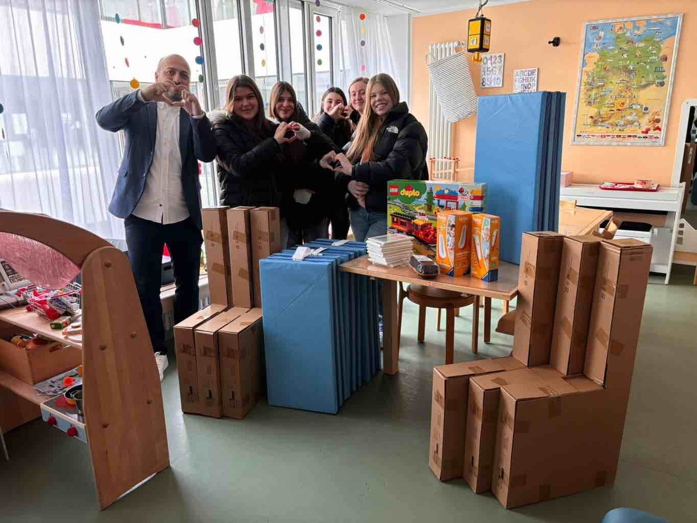 Eine Gruppe von Menschen steht lächelnd in einem Raum voller Spielzeug und Kartons, während sie mit den Händen Herzen formen.