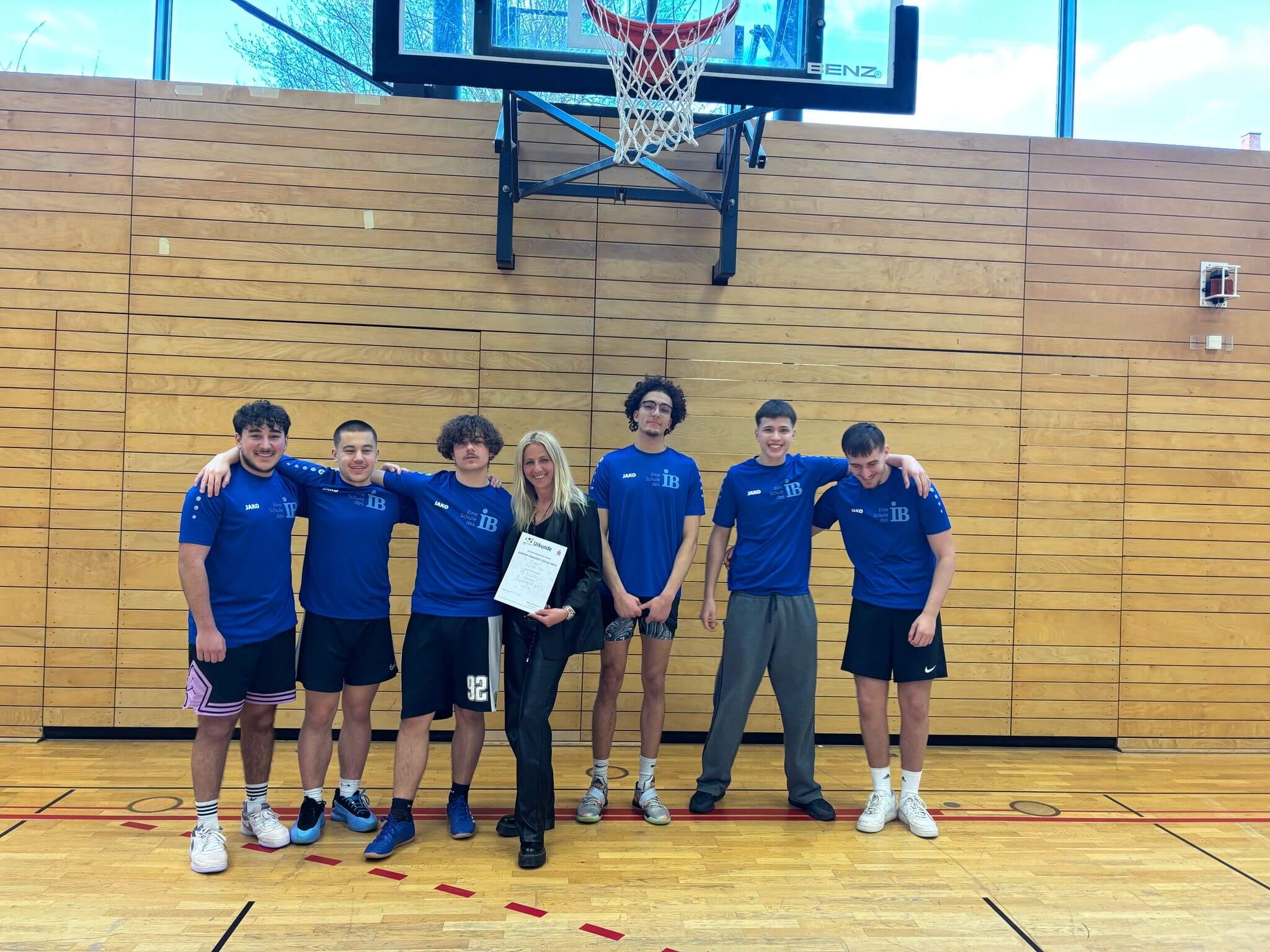 Eine Gruppe von sechs jungen Männern in blauen Trikots posiert lächelnd in einer Sporthalle unter einem Basketballkorb.