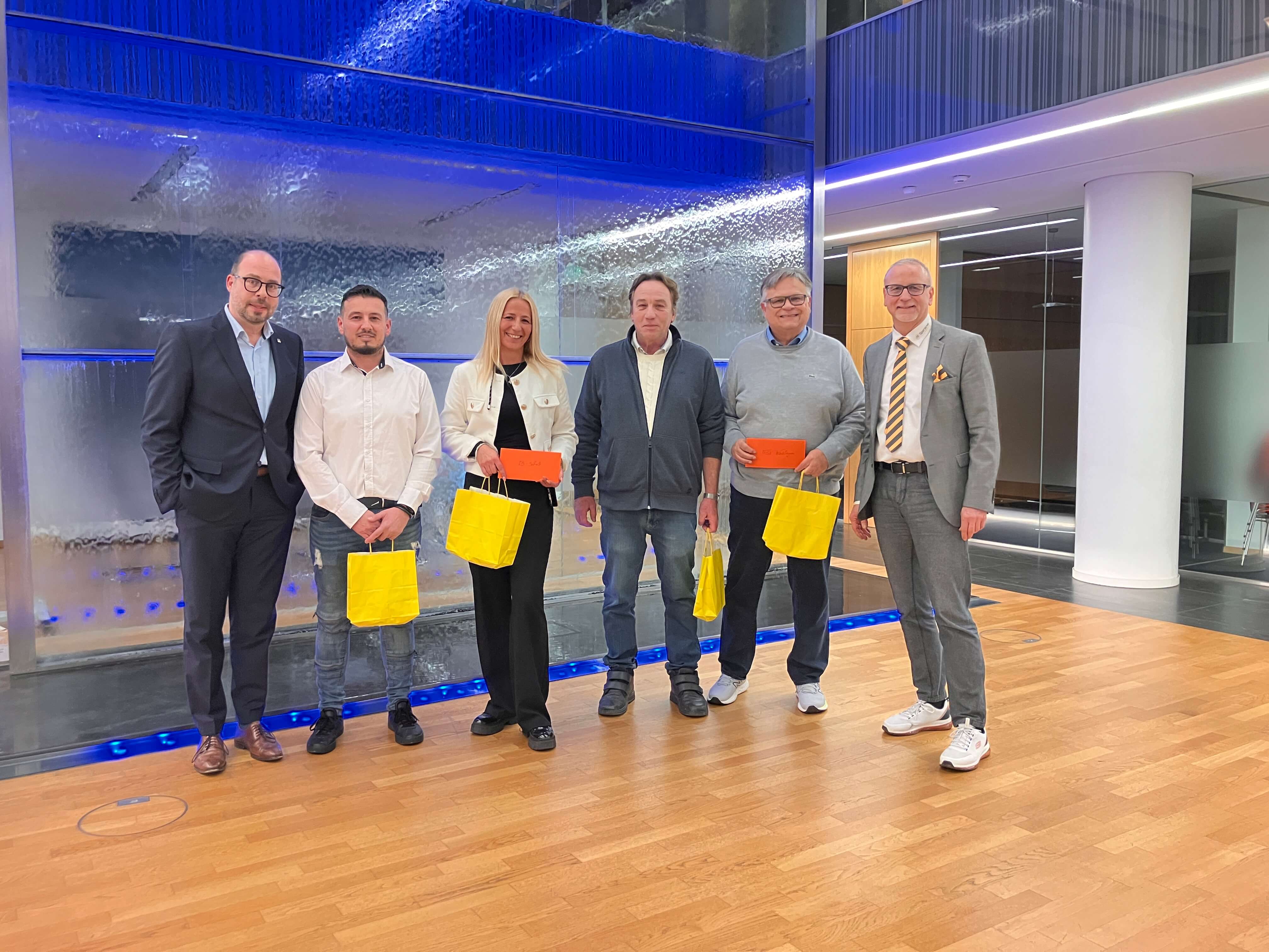 Sechs Personen stehen lächelnd in einem modernen Raum mit blauen Lichtern und tragen gelbe Geschenktüten.
