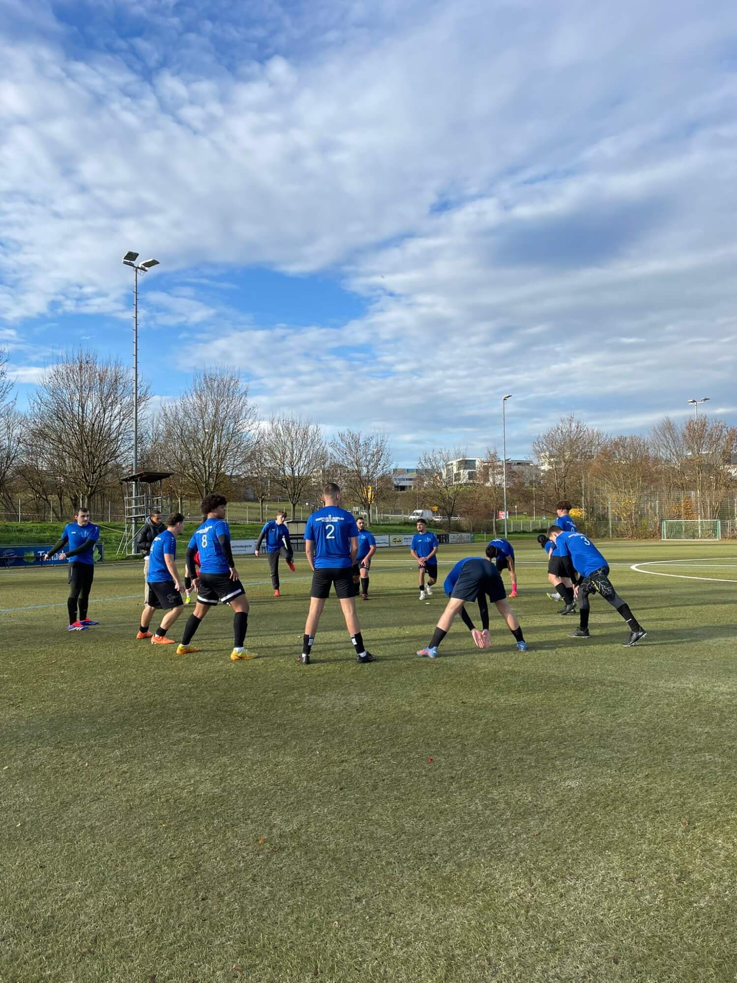 Eine Fußballmannschaft in blauen Trikots dehnt sich auf einem Rasenplatz unter einem bewölkten Himmel vor dem Training.