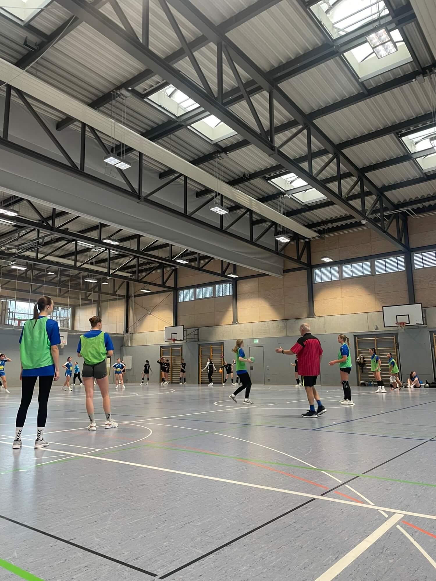 In einer Sporthalle spielen Jugendliche ein intensives Handballspiel unter der Leitung eines Trainers in rotem Trikot.
