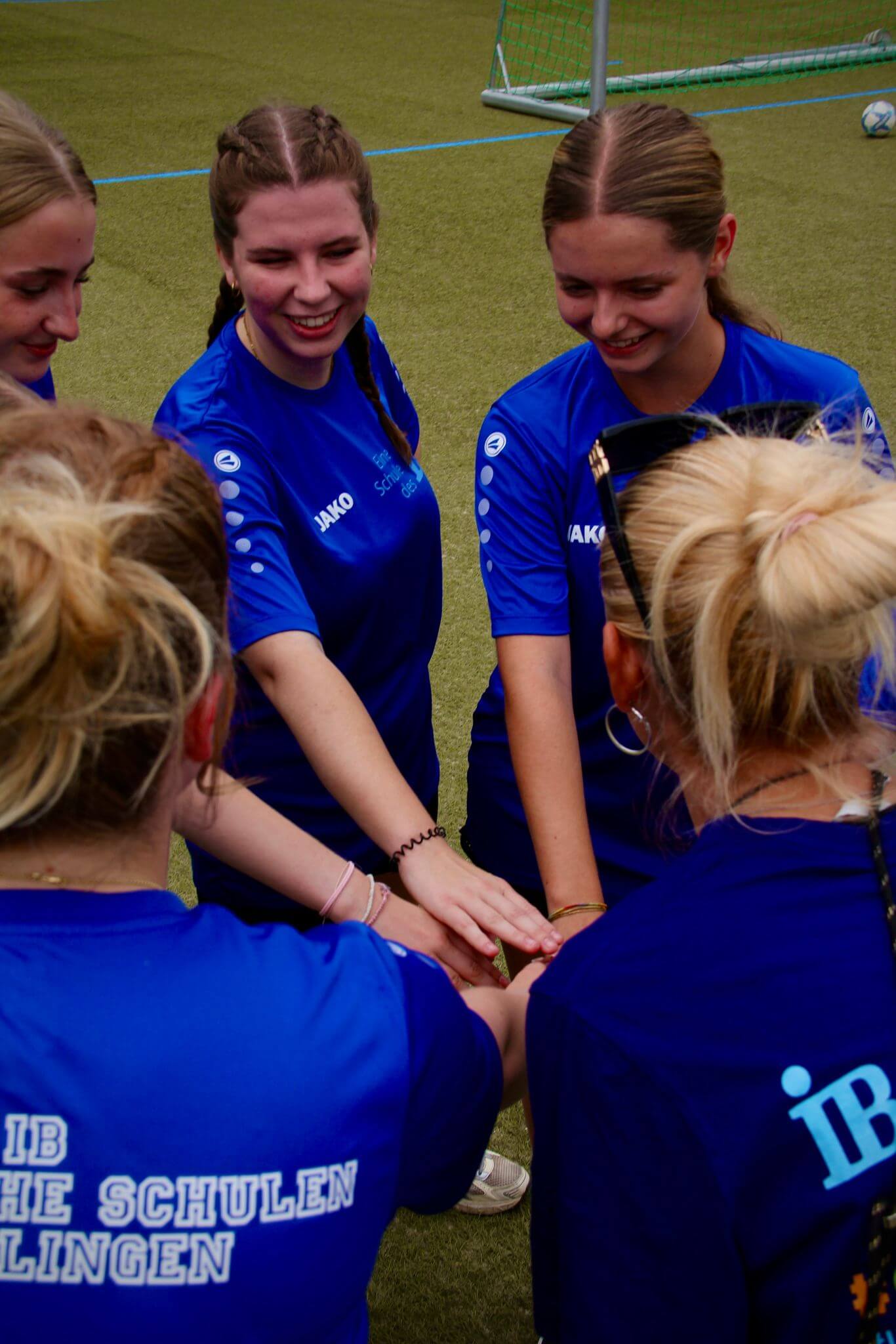 Eine Gruppe junger Frauen in blauen Trikots legt die Hände beim Teamspiel auf einem Sportplatz zusammen.