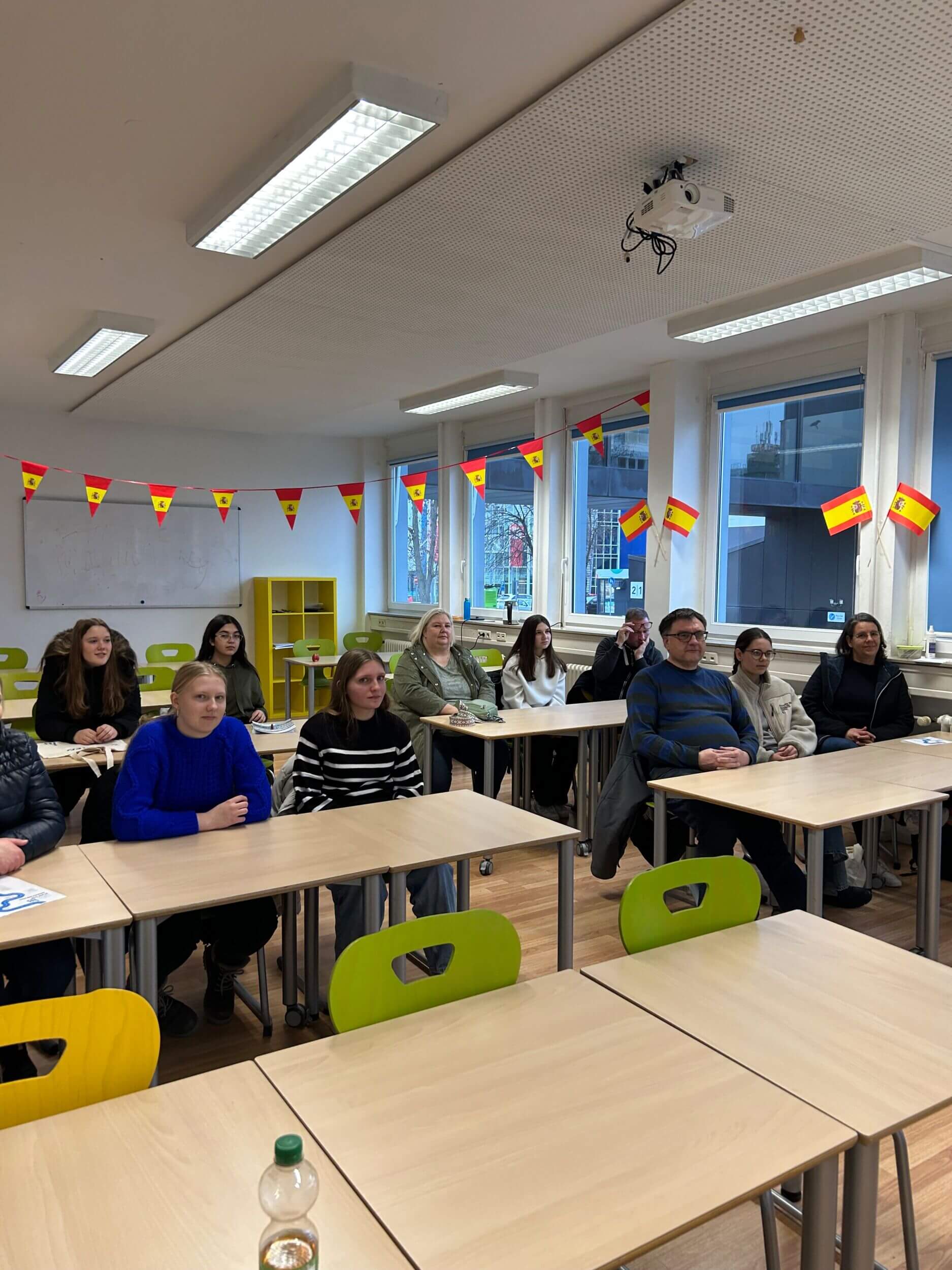 Menschen sitzen in einem Klassenzimmer mit spanischen Fahnen, aufmerksam auf eine Präsentation fokussiert.