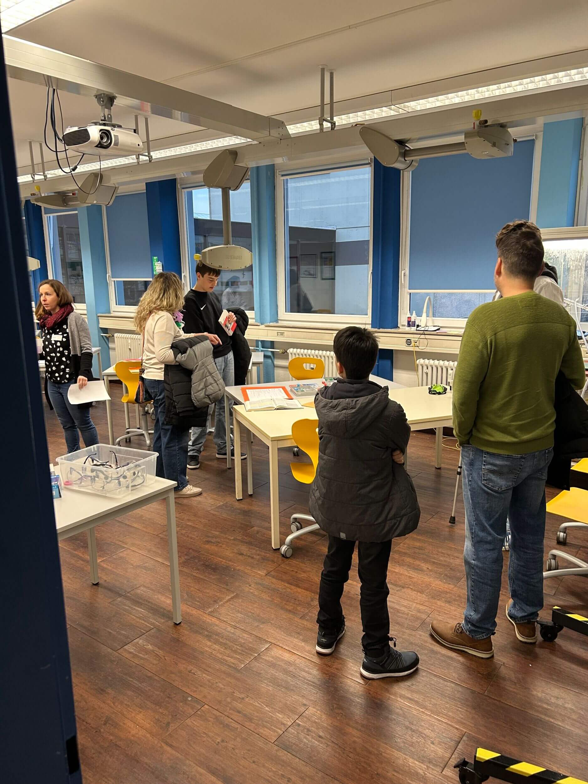 Mehrere Personen stehen in einem Klassenzimmer mit gelben Stühlen und besprechen Schulmaterialien.