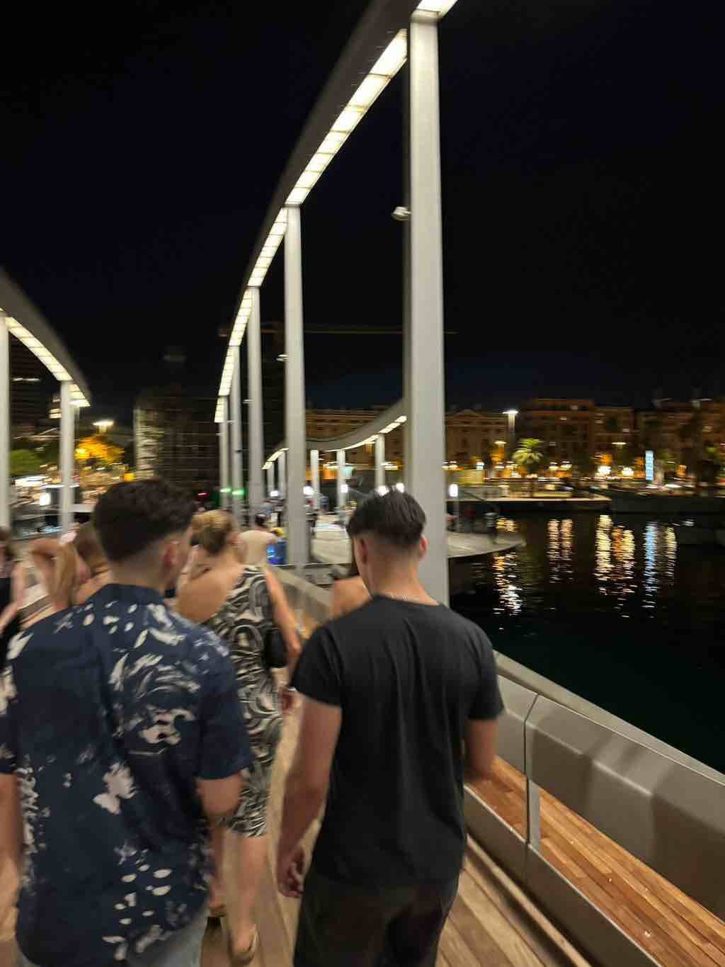Menschen spazieren bei Nacht über eine beleuchtete Brücke in Barcelona mit Blick auf das Wasser.