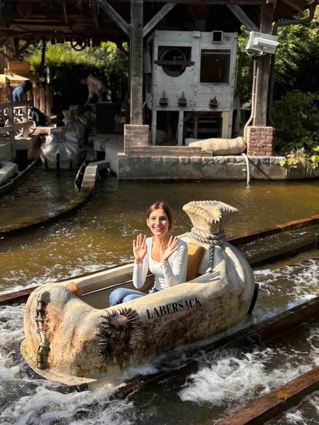 Eine Frau lächelt und winkt aus einem Wasserfahrgeschäft namens Labersack, das durch einen Freizeitpark fährt.